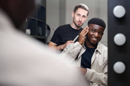 Foto de Peluquero charla con un chico afroamericano, un cliente se sienta frente a un espejo en una silla de peluquero - Imagen libre de derechos