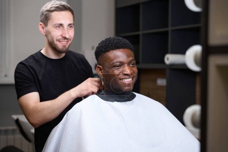 Téléchargez les photos : Barbier avec un bouc et moustache met un voile de protection afro-américain pour un client, l'homme a les cheveux bouclés - en image libre de droit