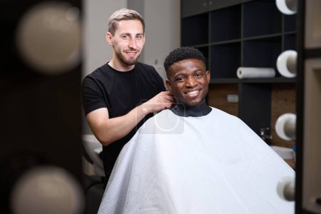 Photo for Hairdresser puts on an African American protective veil for a client, the man has curly hair - Royalty Free Image