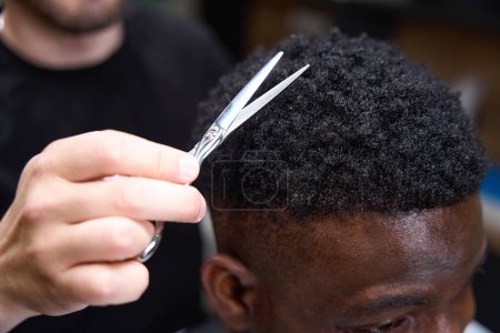 Foto de Peluquería hace un corte de pelo afroamericano para un cliente, un maestro utiliza tijeras - Imagen libre de derechos