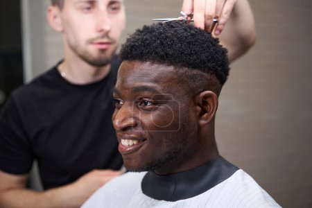 Photo for Young African American guy cuts his hair in a barbershop, the master uses a comb and scissors in his work - Royalty Free Image
