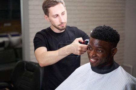 Téléchargez les photos : Coiffeur caucasien fait bordure avec une tondeuse, un client satisfait se trouve dans une chaise de coiffeur - en image libre de droit