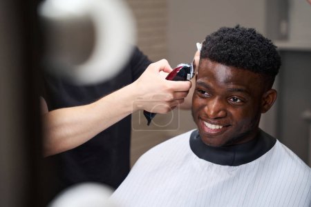 Foto de Sonriente chico afroamericano se sienta en una silla de peluquero, el maestro lo hace bordear con un cortador - Imagen libre de derechos
