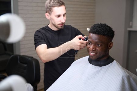 Téléchargez les photos : Beau Afro-Américain assis dans une chaise de coiffeur, le maître le fait bordure avec une tondeuse - en image libre de droit