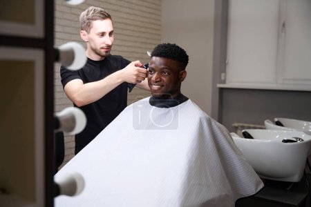 Photo for Master communicates with the client and makes him edging with a clipper, an African American guy in a protective veil - Royalty Free Image