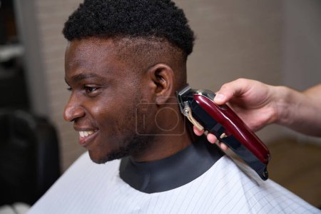 Foto de Un joven afroamericano se corta el pelo en una peluquería, un peluquero usa un clipper en su trabajo - Imagen libre de derechos