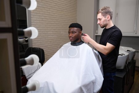Photo for African American guy gets a haircut in a barbershop, the master uses a clipper in his work - Royalty Free Image