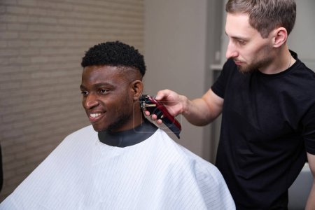 Foto de Alegre afroamericano chico consigue un corte de pelo en una peluquería, un peluquero utiliza un clipper en su trabajo - Imagen libre de derechos