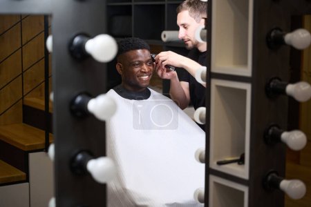Foto de Master corta el pelo de los clientes frente al espejo en la barbería, una escalera de madera se refleja en el espejo - Imagen libre de derechos