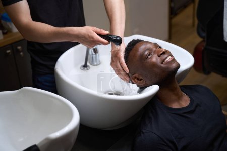 Téléchargez les photos : Barber lave les cheveux d'un gars dans un évier spécial, son client afro-américain a les cheveux bouclés - en image libre de droit