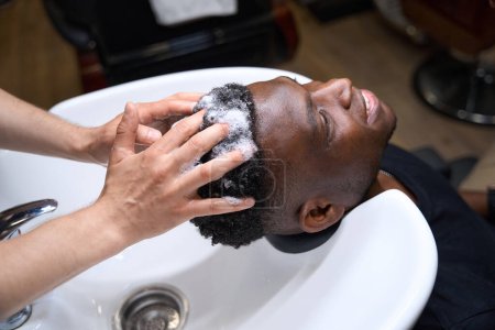 Téléchargez les photos : Homme lave les cheveux des clients avec un shampooing spécial, le mâle afro-américain a les cheveux bouclés - en image libre de droit