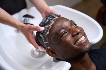 Téléchargez les photos : Satisfait homme afro-américain aime se laver les cheveux dans le salon de coiffure, le coiffeur lave la mousse à l'eau chaude - en image libre de droit