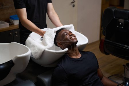 Photo for Caucasian barber dries clients hair with a soft towel, African American guy is comfortably located at a special sink - Royalty Free Image