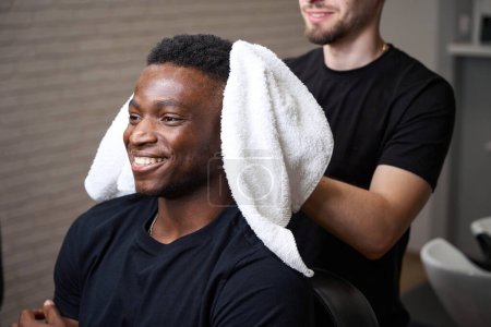 Téléchargez les photos : Jeune coiffeur sèche les cheveux des clients avec une serviette douce, le gars afro-américain s'assoit confortablement dans la chaise de coiffeur - en image libre de droit
