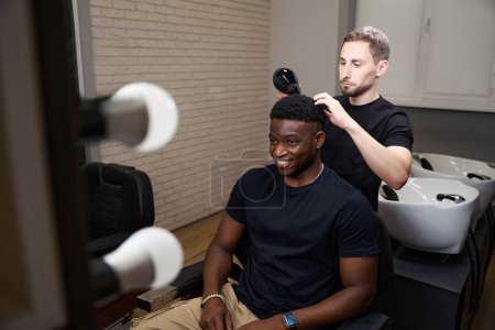 Photo for Barber dries a clients hair with a professional hair dryer, an African American male sits comfortably in a hairdressers chair - Royalty Free Image