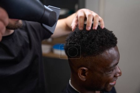 Foto de Estilista seca el pelo rizado de un hombre afroamericano, el maestro utiliza un secador de pelo profesional - Imagen libre de derechos