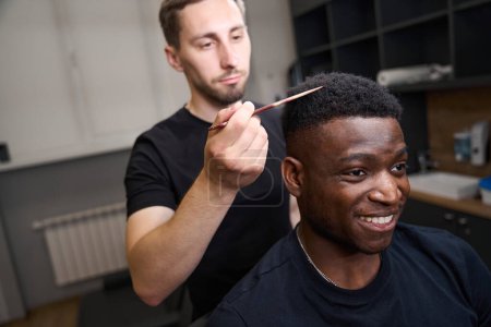 Téléchargez les photos : Styliste coiffant les hommes afro-américains cheveux bouclés, styliste en utilisant un peigne spécial - en image libre de droit