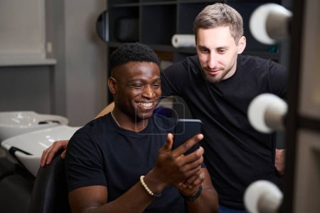 Téléchargez les photos : Satisfait client afro-américain communique en ligne au téléphone dans un salon de coiffure, un coiffeur est debout à proximité - en image libre de droit
