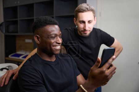 Foto de Sonriente chico afroamericano se comunica en línea por teléfono en una barbería, un barbero está de pie cerca - Imagen libre de derechos
