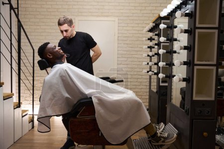 Photo for Stylist works in front of an illuminated mirror as he shaves an African American client with an electric razor - Royalty Free Image