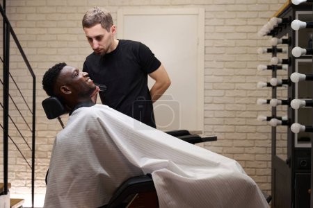 Foto de Estilista afeita a un cliente afroamericano con una afeitadora eléctrica, trabaja delante de un espejo iluminado - Imagen libre de derechos