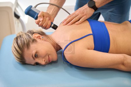 Männlicher Arzt macht Triggerpunkttherapie für Frau, Patientin liegt während extrakorporaler Stoßwellentherapie auf Massagecouch, Rehabilitation