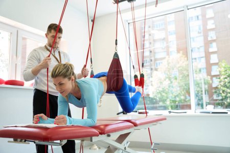 Téléchargez les photos : Femme souriante faisant une thérapie de suspension avec un entraîneur en utilisant le système de cordon rouge dans le centre de remise en forme - en image libre de droit