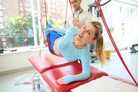 Foto de Mujer feliz mirando a la cámara mientras hace terapia de suspensión con entrenador mediante el uso de cordón rojo en el gimnasio - Imagen libre de derechos