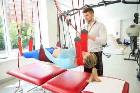 Téléchargez les photos : Femme caucasienne faisant un traitement de suspension avec un entraîneur en utilisant un cordon rouge dans une clinique de fitness - en image libre de droit