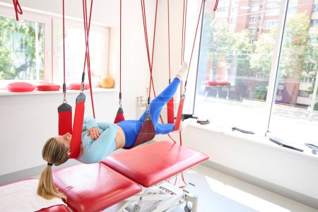 Téléchargez les photos : Jolie femme en tenue de fitness faisant une thérapie de suspension en utilisant le cordon rouge dans la clinique de fitness - en image libre de droit
