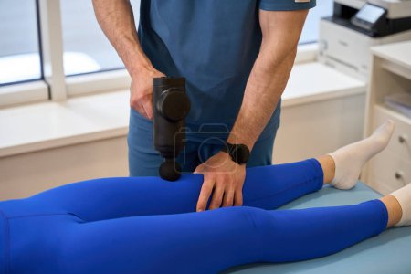 Photo for Male rehabilitologist using special massage gun to reduce muscle stiffness and tension and improve blood flow in the massaged muscles - Royalty Free Image