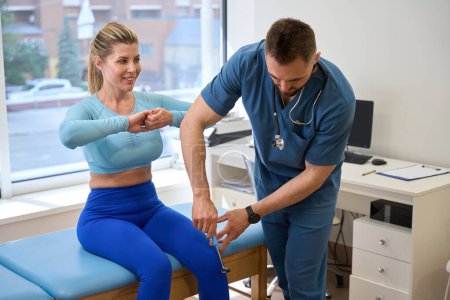 Téléchargez les photos : Femme athlétique regardant beau médecin garder des tests médicaux, en utilisant le marteau pour frapper à genoux et vérifier les réflexes, bilan de santé neurologique, soins médicaux - en image libre de droit
