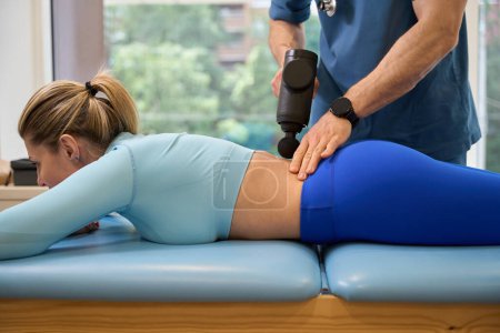Téléchargez les photos : Massothérapeute massant la colonne vertébrale féminine et le bas du dos avec un pistolet de massage, réduisant la rigidité et la tension musculaires et améliorant la circulation sanguine dans les muscles - en image libre de droit