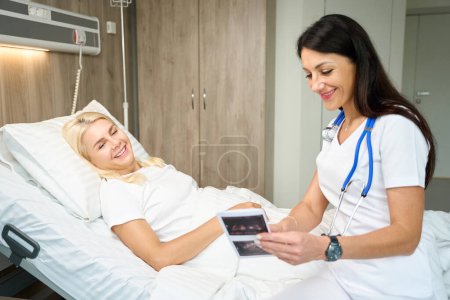 Photo for Happy woman with attending physician reviews ultrasound results, women are in hospital ward - Royalty Free Image