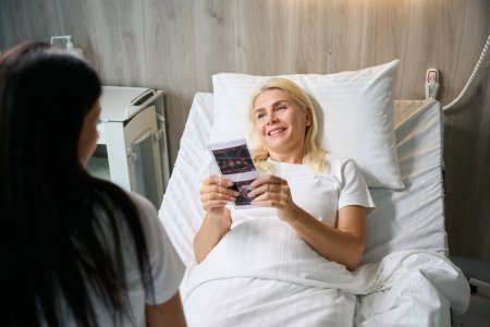 Foto de Mujer feliz sostiene imágenes de ultrasonido en sus manos, se comunica con un médico en una habitación de hospital - Imagen libre de derechos