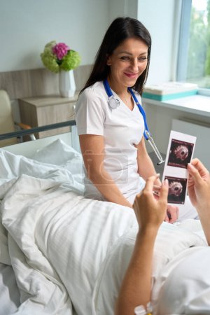Foto de Paciente en una cama de hospital sostiene imágenes de ultrasonido en sus manos, se comunica con un médico en una habitación de hospital - Imagen libre de derechos