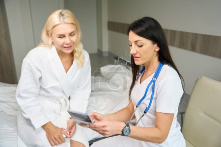 Foto de Dos mujeres están sentadas en una habitación de hospital, un médico y un paciente están mirando imágenes de ultrasonido - Imagen libre de derechos