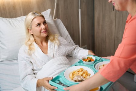 Krankenschwester serviert einer Frau auf dem Krankenhausbett ein Tablett mit Lebensmitteln, der Patient bekommt eine Diät-Mahlzeit