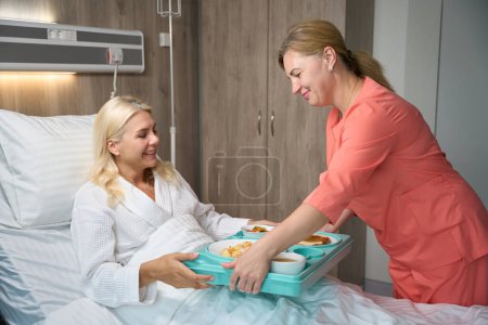 Foto de Amistosa enfermera sirve una bandeja de comida a una mujer en una cama de hospital, el paciente tiene una comida dietética - Imagen libre de derechos