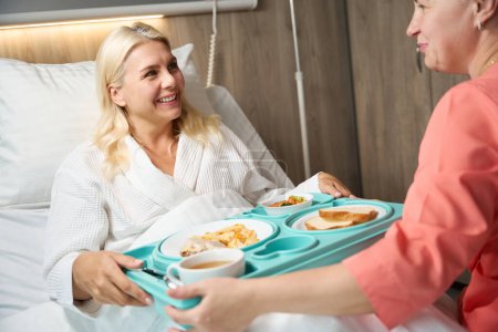 Foto de El empleado del centro médico sirve bandeja de comida a una mujer en una cama de hospital, el paciente tiene una comida dietética - Imagen libre de derechos