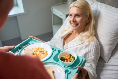 Infirmière prend soin d'une patiente au lit, elle a apporté sa nourriture diététique sur un plateau bleu