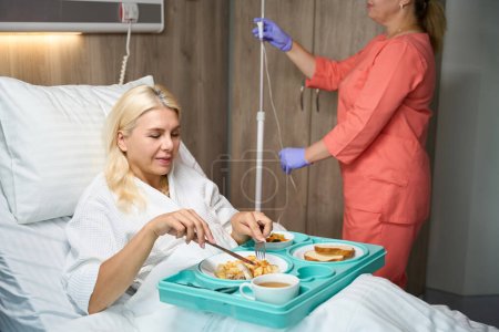 Foto de La paciente en reposo en cama está almorzando en la cama, y una enfermera le está preparando una vía intravenosa. - Imagen libre de derechos