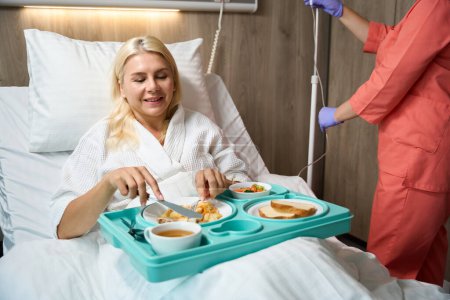 Foto de La mujer en el hospital en reposo en cama está almorzando en la cama, junto a una enfermera preparando una intravenosa para ella. - Imagen libre de derechos