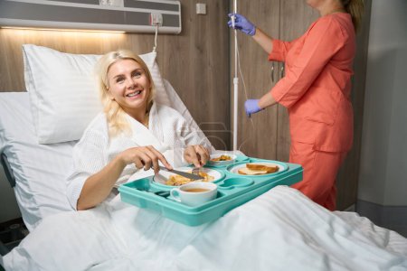 Photo for Recovering patient on bed rest is having lunch in bed, with a nurse nearby preparing an IV for her - Royalty Free Image