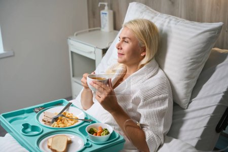 Foto de Paciente en una bata de hospital está desayunando en la cama, ella está en modo hospital - Imagen libre de derechos