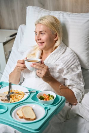 Téléchargez les photos : Récupérer une patiente en blouse d'hôpital prend le petit déjeuner au lit, elle est en mode hôpital - en image libre de droit