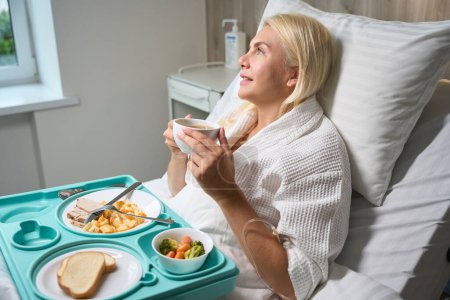 Foto de Recuperar paciente mujer se encuentra en una cama de hospital, delante de ella hay una bandeja azul con comida dietética - Imagen libre de derechos