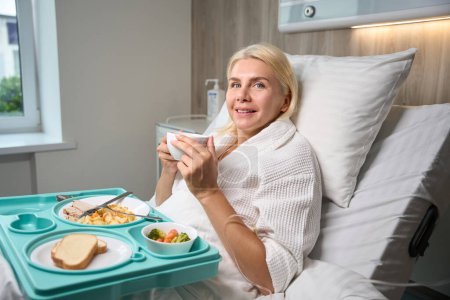 Foto de Hermosa mujer se encuentra en una cama de hospital, delante de ella hay una bandeja azul con comida dietética - Imagen libre de derechos