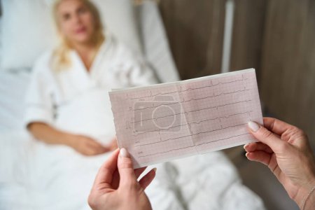 Foto de Médico femenino en una habitación de hospital con resultados de electrocardiograma en las manos, una paciente se acuesta en una cama de hospital - Imagen libre de derechos