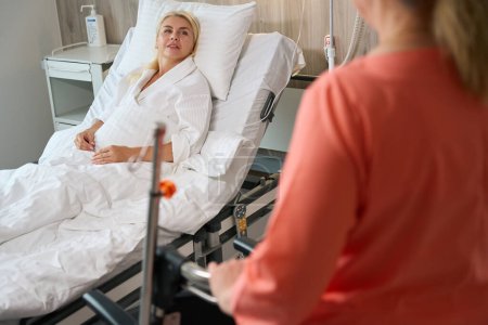 Foto de Una mujer con una bata de hospital se sienta en una cama especial, junto a una enfermera con una silla de ruedas - Imagen libre de derechos